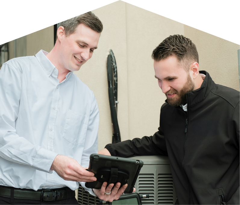 A Romaniuk technician in a white shirt shows service details on a tablet to a smiling client in a black jacket, standing beside an AC unit outside a beige building. A Romaniuk technician in a white shirt shows service details on a tablet to a smiling client in a black jacket, standing beside an AC unit outside a beige building.