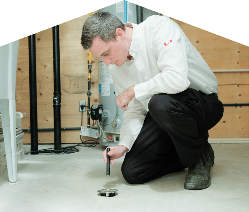 Romaniuk plumbing specialist inspecting a basement floor drain with a flashlight during a drain cleaning service, in a utility room with visible pipes, water heater, and wood panel walls. Romaniuk plumbing specialist inspecting a basement floor drain with a flashlight during a drain cleaning service, in a utility room with visible pipes, water heater, and wood panel walls.