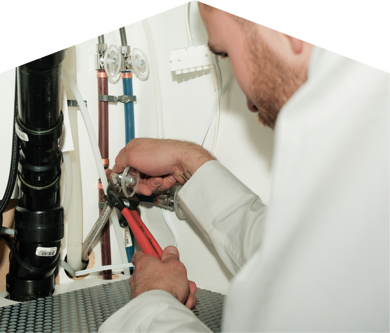 Romaniuk plumbing expert tightening connections under a sink during a faucet installation, surrounded by hot and cold water supply lines and PVC drainage. Romaniuk plumbing expert tightening connections under a sink during a faucet installation, surrounded by hot and cold water supply lines and PVC drainage.