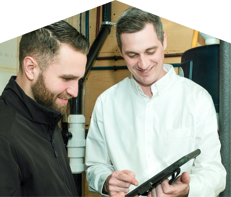 Romaniuk specialist reviewing maintenance plan options with a smiling homeowner using a tablet, inside a utility room with visible piping. Romaniuk specialist reviewing maintenance plan options with a smiling homeowner using a tablet, inside a utility room with visible piping.