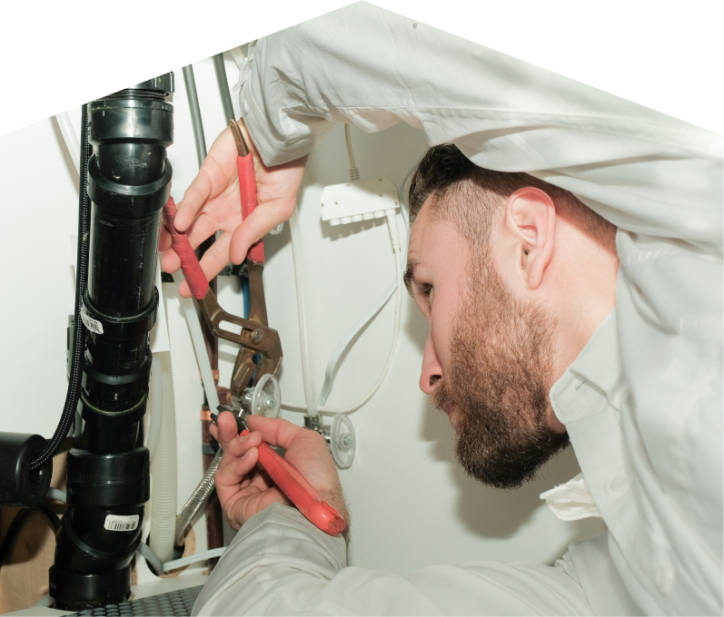 Romaniuk plumbing specialist uses hand tools to tighten pipe fittings beneath a sink during a repair, with visible wiring and drainage lines in the background. Romaniuk plumbing specialist uses hand tools to tighten pipe fittings beneath a sink during a repair, with visible wiring and drainage lines in the background.