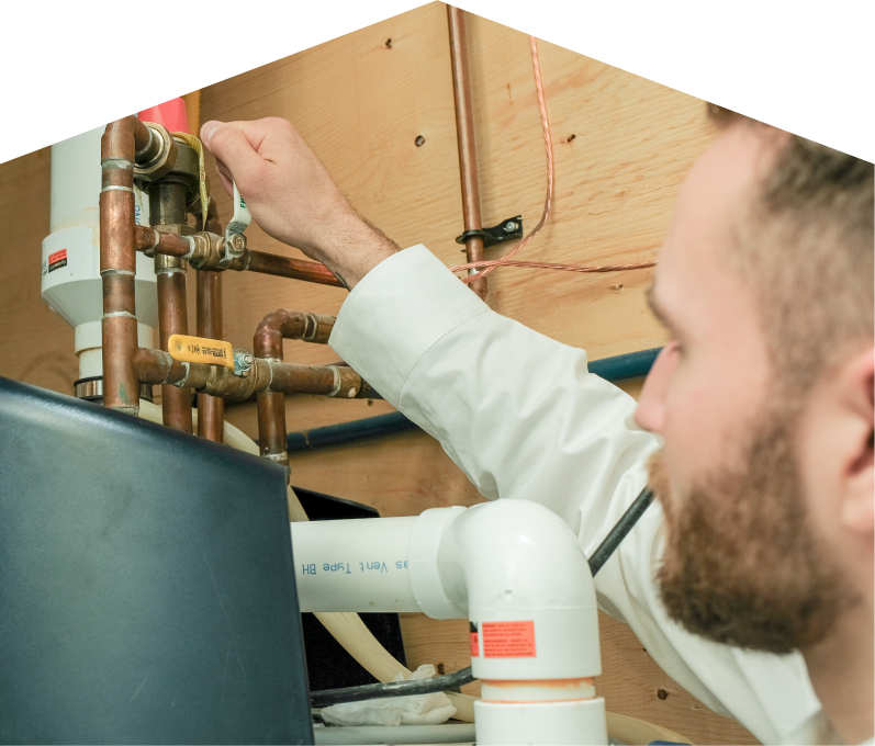 Romaniuk water heater specialist tightens a valve on copper piping during a water heater installation, with exposed wood paneling and wiring in the background. Romaniuk water heater specialist tightens a valve on copper piping during a water heater installation, with exposed wood paneling and wiring in the background.